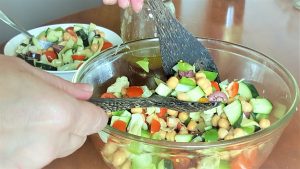 image of tangy greek chickpea salad in a glass bowl being tossed by a pair of wooden spoons. 