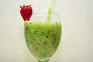 image of glass holding Green Goodness smoothie by intentionally eat for 5 healthy non-dairy smoothies. Green goodness smoothie in a glass with a straw and a strawberry on the lip.