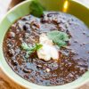 image of vegan black bean soup in a green bowl topped with nondairy sour cream and cilantro with a copper spoon