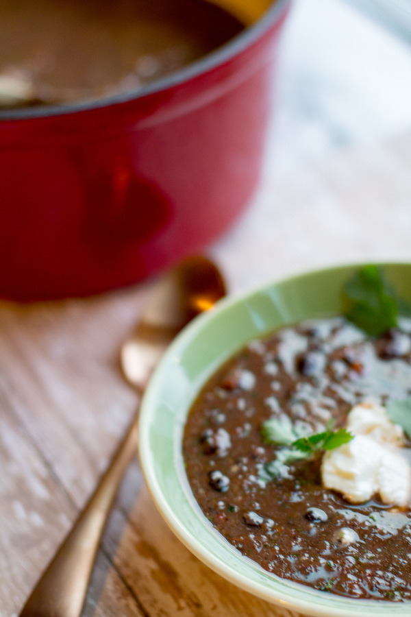image of vegan black bean soup in a green bowl with a copper spoon and red soup pot next to it