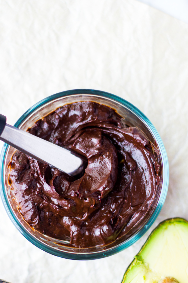 image of chocolate avocado frosting by intentionally eat in a clear glass bowl with a spatula and an avocado next to it