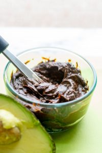 image of chocolate avocado frosting in a clear glass dish with a knife and an avocado next to the dish