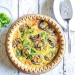 image of vegan quiche with vegetables. Next to it is a small bowl of green onions and a plate with a pie server.