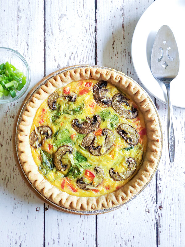 image of vegan quiche with vegetables. Next to it is a small bowl of green onions and a plate with a pie server.