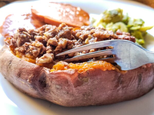 healthy twice baked sweet potato on a white plate being cut by a fork with brussels sprouts and vegan ham in the background