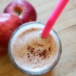 image of a glass full of apple pie smoothie with a pink straw and apples in the background