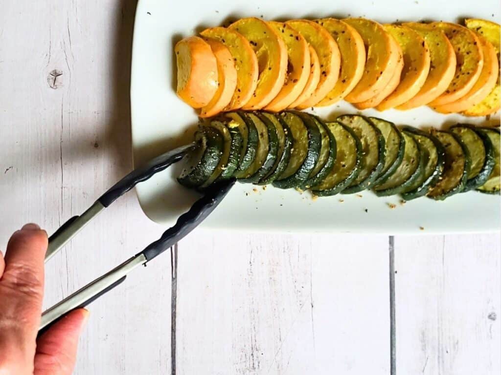 roasted zucchini and squash on a white plate with a hand and tongs