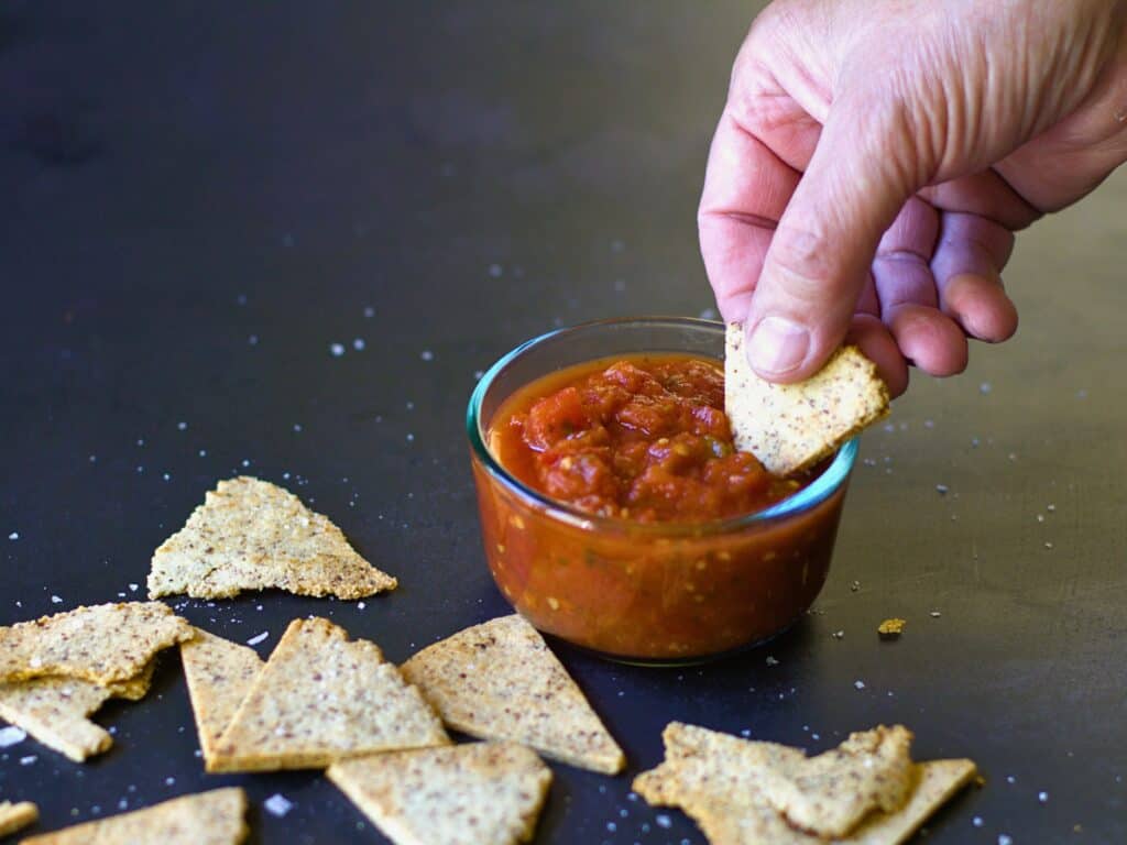 a hand dipping a keto tortilla chip into salsa