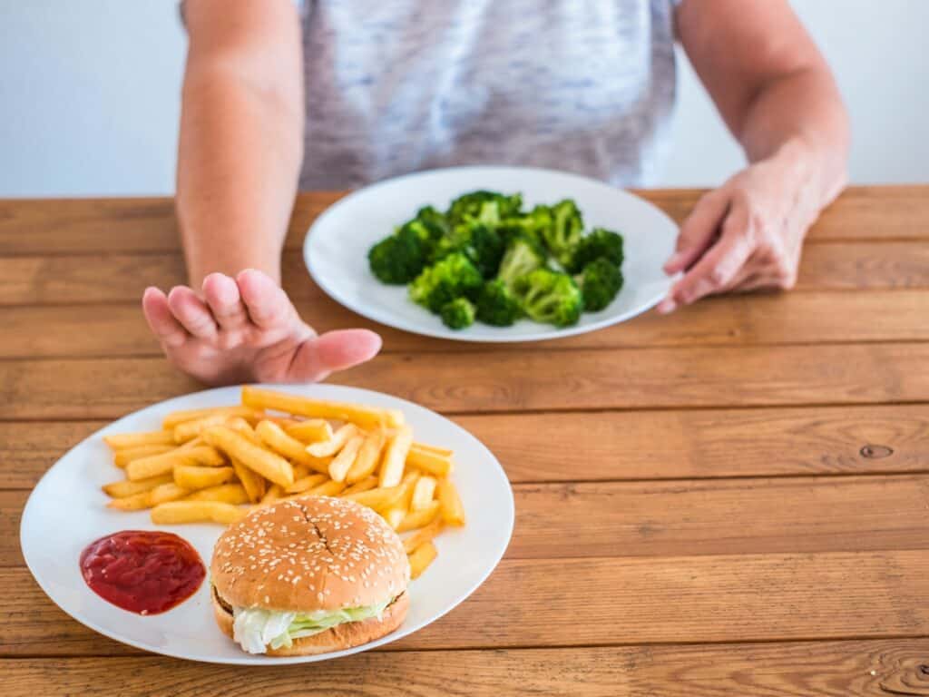 a person holding a plate of broccoli and pushing away a plate with a burger and fries