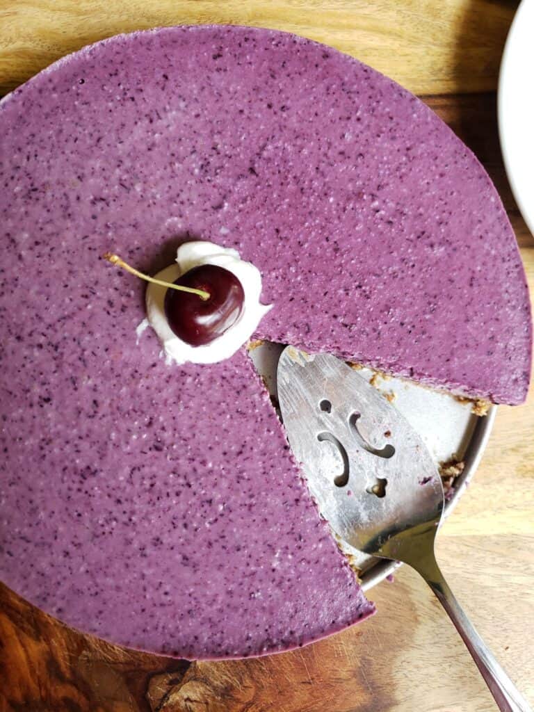 Slice of high-protein vegan blueberry cheesecake on a plate, dairy-free with a date-oat crust and cocoa whip on top.